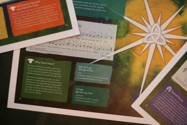 Several pages from the Gospel Story Hymnal are spread out on the table. Image is zoomed in on the 'blurbs,' short notes about the hymns. Visible blurbs are titled "Why start here?," "All Creation Reveals and Glorifies God," "God of Power," and the definitions for "doxology" and "amen."
