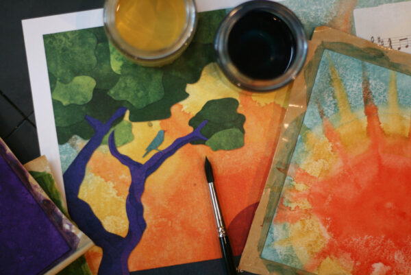 A page from the Gospel Story Hymnal is on the table along with watercolored pages, paints, and a paintbrush. The illustration shows a bird sitting on a tree branch.