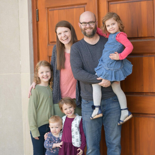 Lindsey standing with her family in front of a large door. She has a husband and four young daughters.